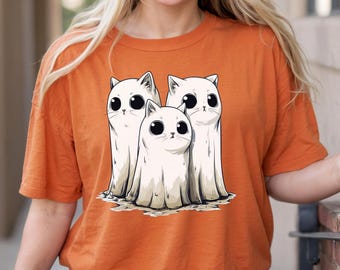 以下が含まれることがあります： オレンジ色のTシャツ。3匹の白い猫の幽霊のグラフィックが描かれています。大きな黒い目をした猫たちは、漫画風のシンプルなデザインです。Tシャツは無地のオレンジ色です。