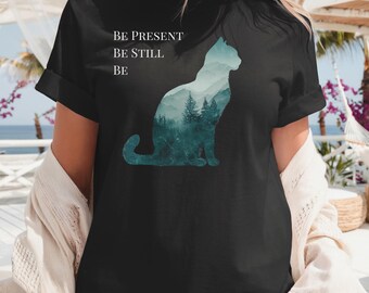 May include: Black t-shirt featuring a cat silhouette filled with a forest scene in shades of teal and green. The shirt has the words "BE PRESENT, BE STILL, BE" printed in white. The shirt has rolled-up sleeves.