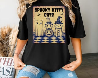 以下が含まれることがあります： ハロウィーンをテーマにしたグラフィックが特徴の黒いTシャツ。「Spooky Kitty Cats」の文字と、幽霊、カボチャの頭の猫、魔女の帽子をかぶった猫のイラストが描かれています。コウモリも含まれています。