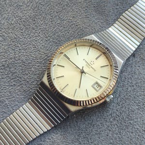 May include: A vintage Eterna-matic 1000 wristwatch with a silver-tone metal band and a cream-colored face. The watch has a gold-tone bezel and a date display at the 3 o'clock position. The watch is displayed on a gray surface.