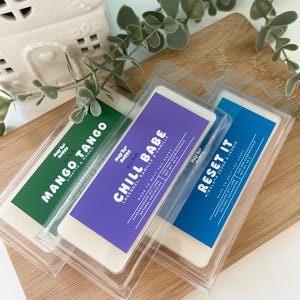 Könnte beinhalten: Drei Wachsduftriegel in durchsichtiger Kunststoffverpackung, jeweils mit einem unterschiedlich farbigen Etikett. Die Etiketten lauten "Mango Tango", "Chill Babe" und "Reset It". Die Riegel liegen auf einem Holzschneidebrett mit Grünzeug im Hintergrund.