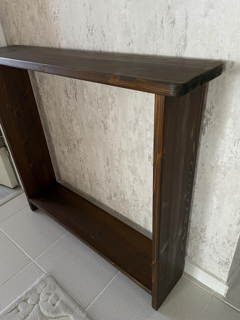 May include: A dark brown wooden console table with a rectangular top and a lower shelf. The table has a simple, open design and is made of solid wood. The finish is a rich, dark stain, and the table is approximately 122 cm long.
