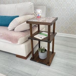 May include: A three-tiered, dark-stained wooden side table. The top surface features a decorative tile design. A mug with a cat illustration sits on top. A small potted cactus and a book are on the lower shelves.