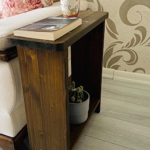 May include: A dark brown wooden side table with a lower shelf, holding a small potted cactus. A book and a decorative mug sit on top. The table is next to a floral patterned pillow.