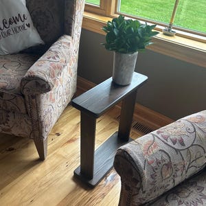 Puede incluir: Una mesa auxiliar de madera marrón oscuro con una parte superior rectangular y dos patas de soporte. Una pequeña maceta cilíndrica gris con plantas de hojas verdes se encuentra encima de la mesa. La mesa está colocada entre dos sillones estampados.