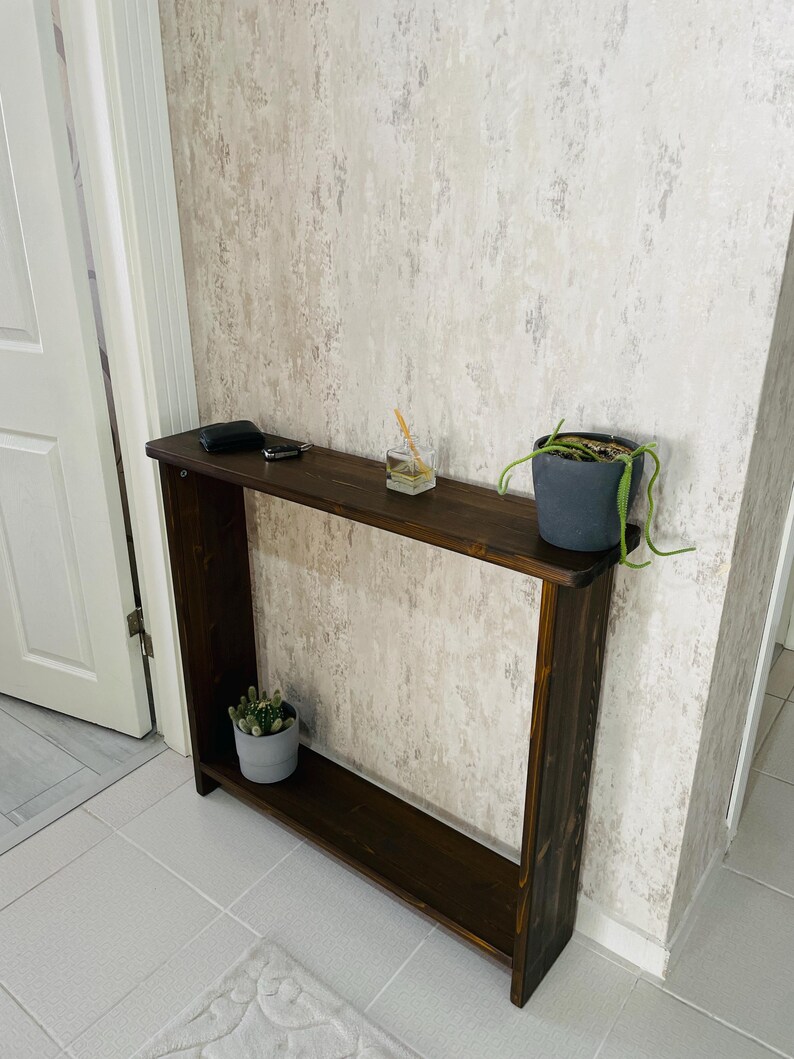 May include: A dark brown wooden console table with a rectangular design and a lower shelf. A small potted cactus and a small potted plant with trailing vines sit on the table. A small black object and a key are on the top shelf.