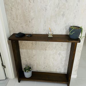 May include: A dark brown wooden console table with a lower shelf. A small potted plant, a wallet, keys, and a diffuser sit on the table. The table is against a textured, light-coloured wall.