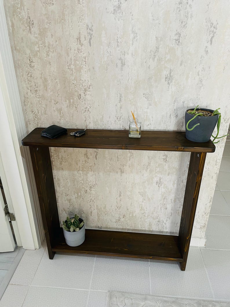 May include: A dark brown wooden console table with a lower shelf. A small potted plant, a wallet, keys, and a diffuser sit on the table. The table is against a textured, light-coloured wall.
