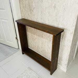 May include: A dark brown wooden console table with a rectangular top and open sides. The table has a lower shelf and is placed against a textured wall. The table is approximately 122 cm long.