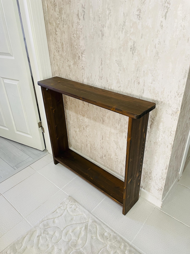 May include: A dark brown wooden console table with a rectangular top and open sides. The table has a lower shelf and is placed against a textured wall. The table is approximately 122 cm long.