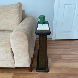 Peut inclure: Une table d'appoint en bois brun foncé avec un plateau rectangulaire et un seul support vertical. Un mug vert et un livre reposent sur la surface de la table. La table est placée à côté d'un canapé beige.
