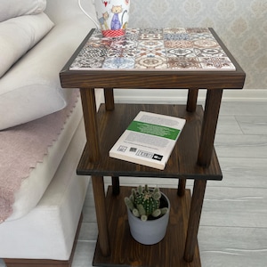 May include: A dark brown, three-tiered side table with a tiled top. A mug with a cat design sits on top. A book and a small potted cactus are on the lower shelves. The table is made of wood and has a simple, rustic design.
