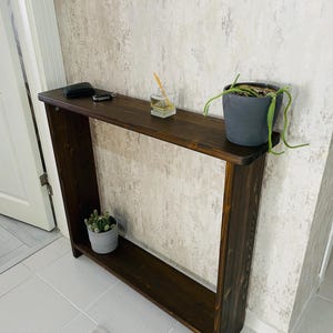 Könnte beinhalten: Eine dunkelbraune Holzkonsolentisch mit einem unteren Regal. Der Tisch steht vor einer strukturierten Wand. Kleine Topfpflanzen und Accessoires sind auf dem Tisch ausgestellt. Der Tisch ist etwa 120 cm lang.