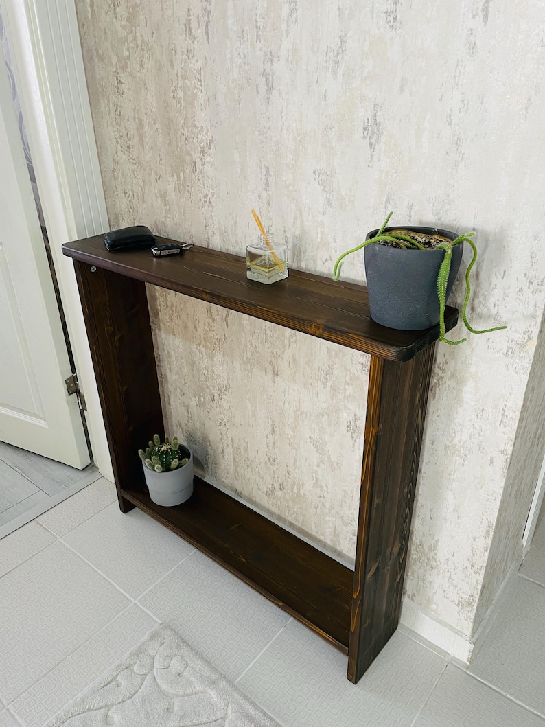 May include: A dark brown wooden console table with a lower shelf. The table is against a textured wall. Small potted plants and accessories are displayed on the table. The table's dimensions are approximately 120 cm in length.
