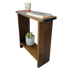 May include: A dark brown wooden side table with a lower shelf. A white mug with a red and white design and a book rest on top. A small potted cactus sits on the lower shelf. The table is made of solid wood.