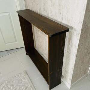 May include: A dark brown wooden console table. The rectangular table has a long, flat top and a lower shelf, with open sides. The wood grain is visible. The table is positioned against a wall.