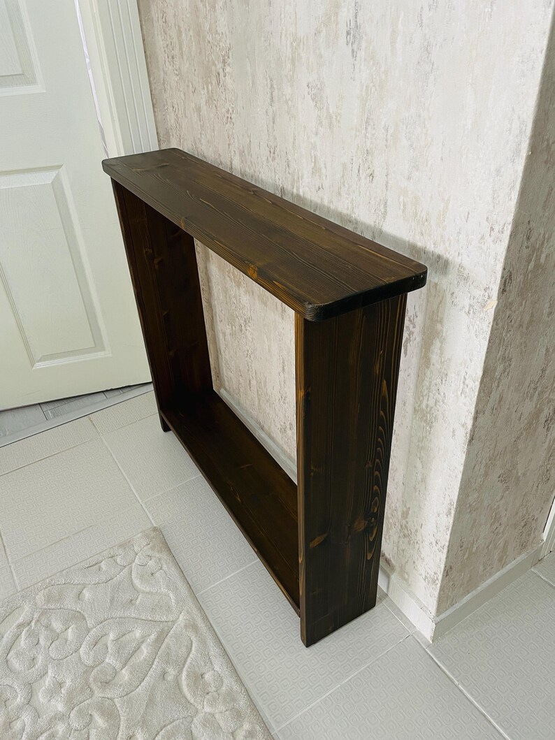 May include: A dark brown wooden console table. The rectangular table has a long, flat top and a lower shelf, with open sides. The wood grain is visible. The table is positioned against a wall.