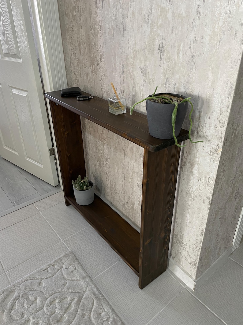 May include: A dark brown wooden console table with a lower shelf. A small potted cactus and a plant in a gray pot sit on the table. A small glass container and keys are also on the table.