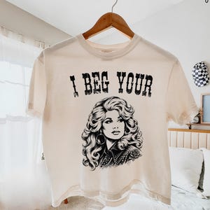 May include: An ivory short-sleeve t-shirt featuring a black graphic print of a person with voluminous hair and the text I BEG YOUR printed in a distressed font above the illustration. The shirt is displayed on a wooden hanger against a light background.
