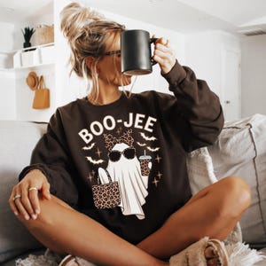 May include: A dark brown crewneck sweatshirt featuring a graphic of a white ghost wearing leopard print sunglasses, a matching bow, and carrying a leopard print tote bag. The ghost holds a coffee cup, surrounded by bats and the text BOO-JEE.