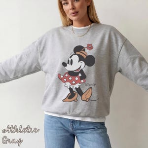 K&ouml;nnte beinhalten: Graues Sweatshirt mit Minnie Maus-Grafik. Die Grafik zeigt Minnie Maus in einem roten Polka-Dot-Rock, braunen Schuhen und einer Blume auf ihrem Hut. Das Wort "Disney" ist darunter aufgedruckt. Der Text "Athletic Gray" ist unten links aufgedruckt.