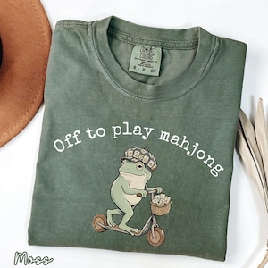 May include: Moss green t-shirt with the phrase "Off to play mahjong" in white. A cartoon frog wearing a hat and riding a scooter is featured below the text. The frog has a basket with mahjong tiles. The shirt is labeled "Comfort Colors".