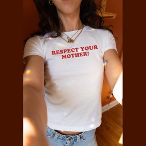May include: A white short-sleeved t-shirt featuring the text "RESPECT YOUR MOTHER!" printed in bold red capital letters across the chest. The shirt is paired with light blue denim jeans, and the overall look is accessorized with gold necklaces.