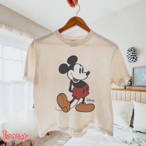 May include: Ivory-colored t-shirt featuring a classic Mickey Mouse graphic. The cartoon character is depicted in black, red, and brown, with the Disney logo. The shirt is hanging on a wooden hanger.
