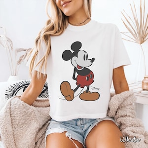 May include: White t-shirt featuring a classic Mickey Mouse graphic. The cartoon character is depicted in his iconic pose, wearing red shorts and brown shoes. The Disney logo is visible. The word "White" is printed at the bottom.