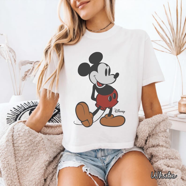 May include: White t-shirt featuring a classic Mickey Mouse graphic. The cartoon character is depicted in his iconic pose, wearing red shorts and brown shoes. The Disney logo is visible. The word "White" is printed at the bottom.