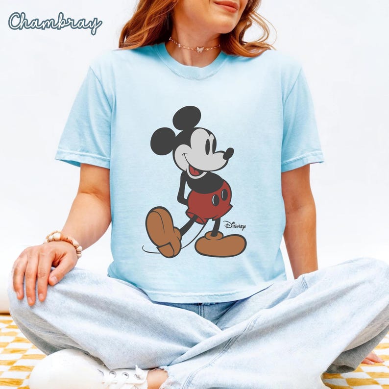 May include: Light blue t-shirt featuring a graphic of Mickey Mouse in his classic pose. The cartoon character is depicted in black, white, red, and brown. The word "Disney" is printed below the character. The word "Chambray" is printed in the upper left corner.