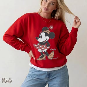 K&ouml;nnte beinhalten: Ein rotes Sweatshirt mit einem Minnie Mouse-Motiv. Die Grafik zeigt Minnie Mouse in einem gepunkteten Kleid und Hut. Das Wort "Disney" ist unter der Figur aufgedruckt. Das Wort "Red" ist unten links aufgedruckt.