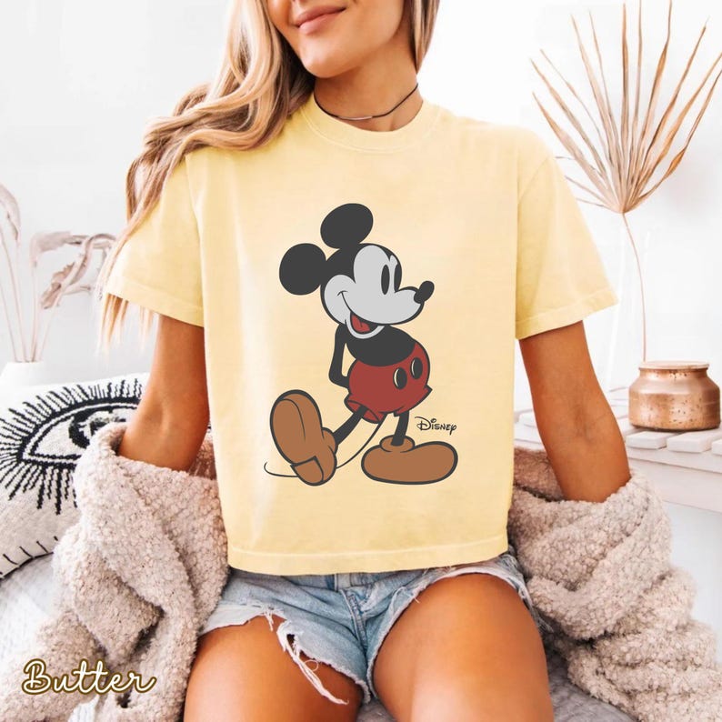 May include: Yellow t-shirt featuring a classic Mickey Mouse graphic. The cartoon character is depicted in his iconic red shorts, brown shoes, and black and white features. The Disney logo is visible. The word "Butter" is printed on the bottom left.