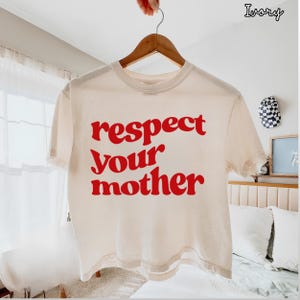 May include: An ivory short-sleeve t-shirt featuring the red text "respect your mother" printed in a bold, wavy font across the center of the chest. The garment is displayed on a wooden hanger against a bright, neutral-toned indoor setting.