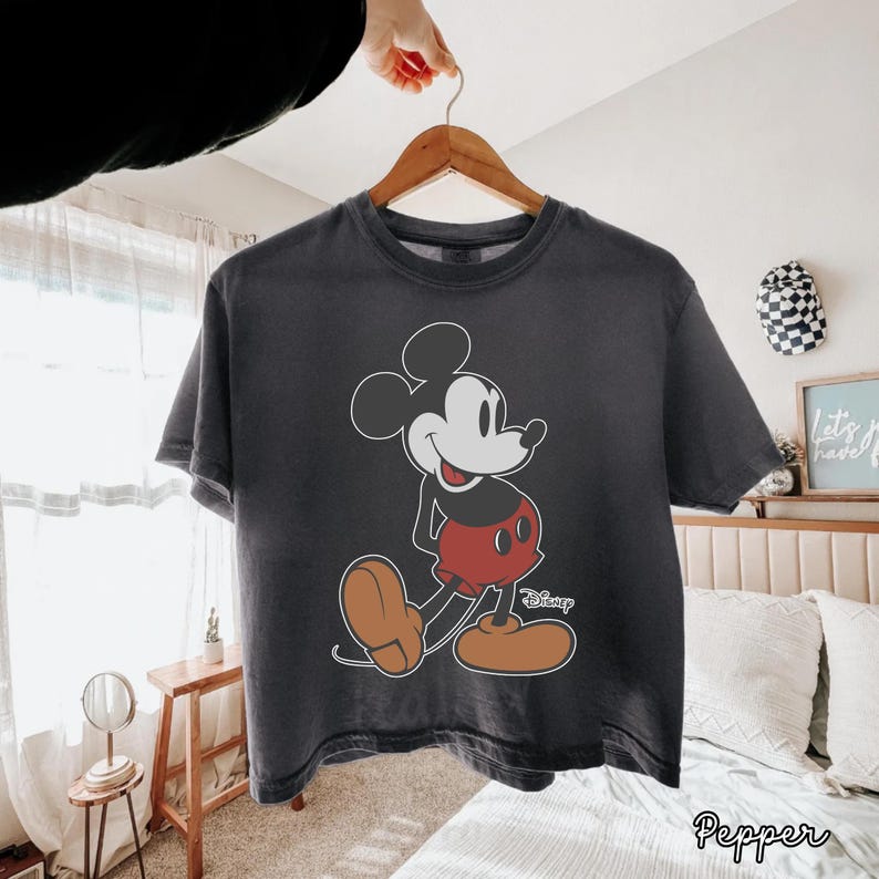 May include: Dark gray t-shirt featuring a classic Mickey Mouse graphic. The cartoon character is depicted in his iconic pose, wearing red shorts and brown shoes. The shirt is hanging on a wooden hanger.