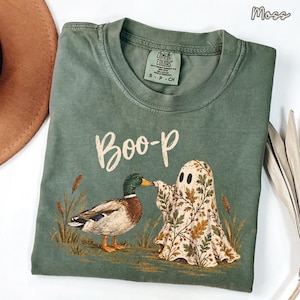 May include: A moss green short-sleeve t-shirt featuring a graphic of a mallard duck and a ghost covered in an autumn leaf pattern. The text "Boo-p" is printed above the figures. The shirt has a Comfort Colors label at the inner neckline.