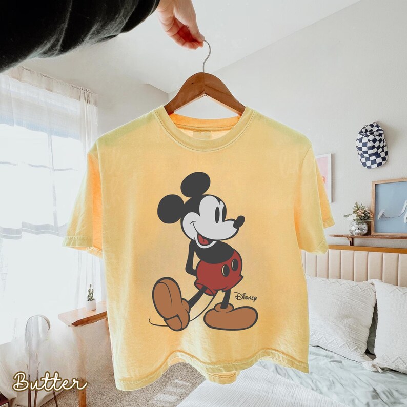 May include: A yellow t-shirt featuring a graphic of Mickey Mouse in a walking pose. Mickey is depicted with black ears, a white face, red shorts, and brown shoes. The Disney logo is printed on the lower right. The word "Butter" is printed on the lower left.
