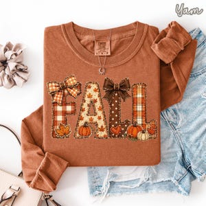 May include: A rust-colored long-sleeve t-shirt featuring the word FALL in large, decorative letters. Each letter has a unique pattern including plaid, autumn leaves, and polka dots, accented with bows and small pumpkin illustrations.