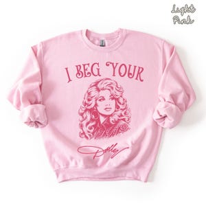 May include: A light pink crewneck sweatshirt featuring a dark pink graphic print of a woman with voluminous hair. The text "I BEG YOUR" is printed above the portrait, and the signature "Dolly" is printed below. The words "Light Pink" appear in the corner.