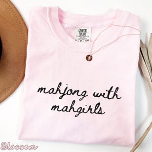 以下が含まれることがあります： 「mahjong with mahgirls」の黒い筆記体で書かれたライトピンクのTシャツ。ペンダント付きのローズゴールドのネックレスがシャツの上にかけられています。茶色の帽子が左上に部分的に見えています。