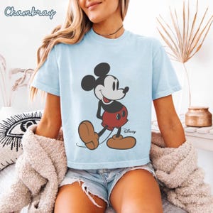 May include: Light blue t-shirt featuring a classic Mickey Mouse graphic. The cartoon character is depicted in his iconic red shorts, brown shoes, and black ears. The Disney logo is printed below the character. The word "Chambray" is printed at the top left.