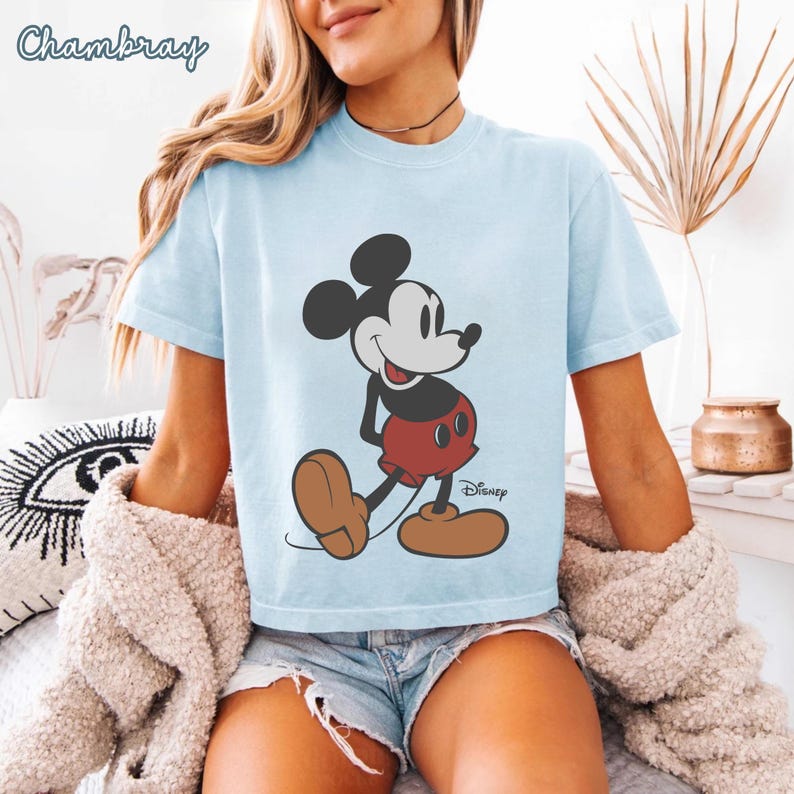 May include: Light blue t-shirt featuring a classic Mickey Mouse graphic. The cartoon character is depicted in his iconic red shorts, brown shoes, and black ears. The Disney logo is printed below the character. The word "Chambray" is printed at the top left.