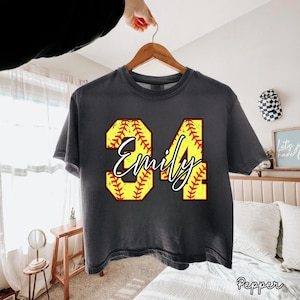 Op de afbeelding: Een donkergrijs t-shirt met een softbalthema. Het ontwerp toont het nummer 34 in geel met rode stiksels en de naam "Emily" in een wit script. Het shirt hangt aan een houten hanger.