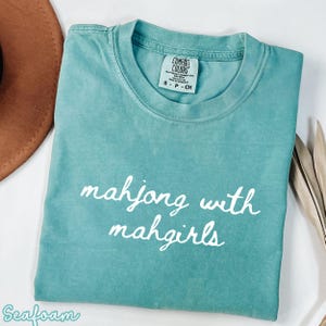 Könnte beinhalten: Gefaltetes, meergrünes T-Shirt mit dem weißen Schriftzug "mahjong with mahgirls". Das Shirt hat ein Comfort Colors-Etikett. Ein brauner Hut und getrocknete Blätter sind im Hintergrund.