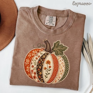 May include: An espresso-coloured t-shirt featuring a large, central pumpkin graphic. The pumpkin is composed of various patchwork-style fabric segments in orange, brown, and green floral and plaid patterns, with a dark green leaf and brown stem.