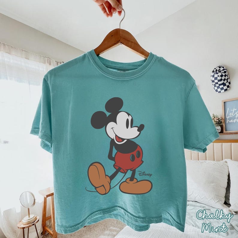 May include: A teal-colored t-shirt featuring a classic Mickey Mouse graphic. The cartoon character is depicted in black, red, and tan, with the Disney logo printed below. The shirt is hanging on a wooden hanger.