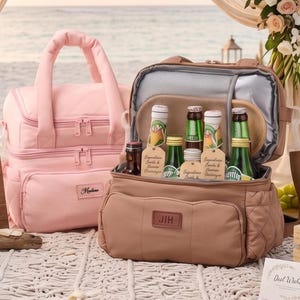 以下が含まれることがあります： ピンクとタンの2つの断熱クーラーバッグがビーチに展示されています。ピンクのバッグには上部のハンドルとフロントポケットが付いています。タンのバッグは開いており、飲み物のボトルが明らかになっています。イニシャルと結婚式の日付が書かれた木製の看板が前景にあります。
