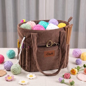 May include: A brown corduroy craft tote bag filled with colorful yarn balls, featuring exterior pockets holding crochet hooks and knitting needles, a zippered front compartment with gold scissors, and a brown leather patch labeled Linda.