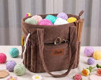May include: A brown corduroy craft tote bag filled with colourful yarn balls, featuring exterior pockets holding crochet hooks and knitting needles, a zippered front compartment with gold scissors, and a brown leather patch labelled Linda.