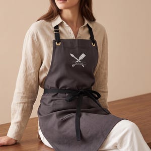 May include: A charcoal gray apron featuring black adjustable neck straps with gold-tone metal hardware. The chest area displays an embroidered white graphic of two crossed paintbrushes above the name Severt, which is underlined by a horizontal line.
